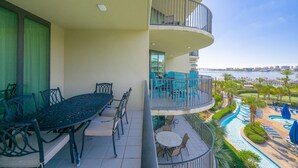 A balcony with a dining table and chairs, a small round table with chairs, and a view of a resort area with a pool and palm trees.