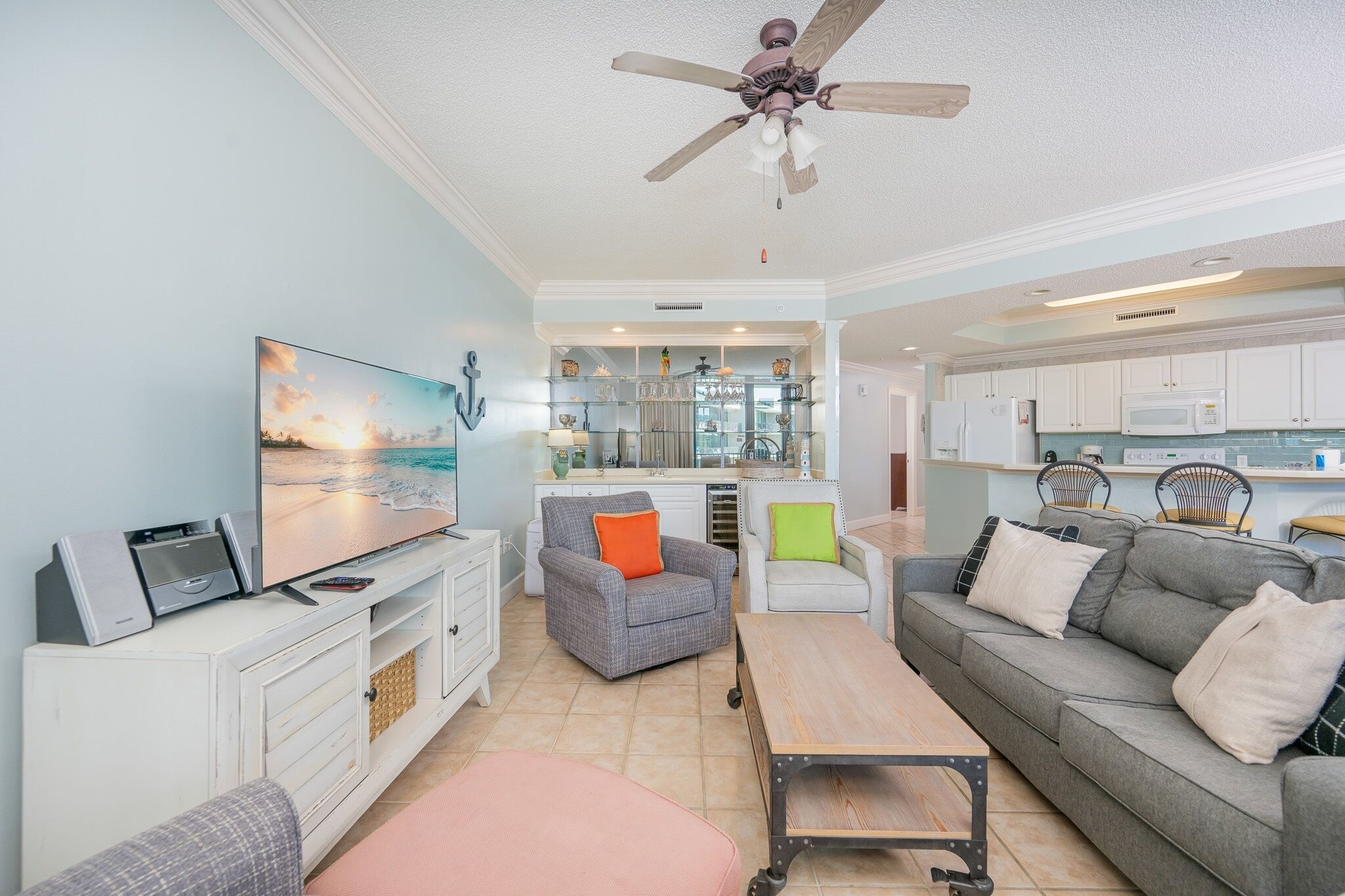 A modern living room with a flat-screen TV, a ceiling fan, and a kitchen area with white cabinets and a microwave.