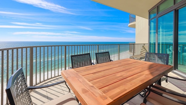 A balcony with a wooden table and chairs, overlooking a beach and ocean. - Phoenix West 1606 3 Bedroom Condo (Orange Beach)