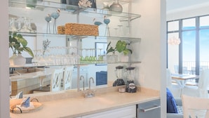 A kitchen with glass shelves displaying glassware, a blender, and a sink. - Phoenix West 1606 3 Bedroom Condo (Orange Beach)