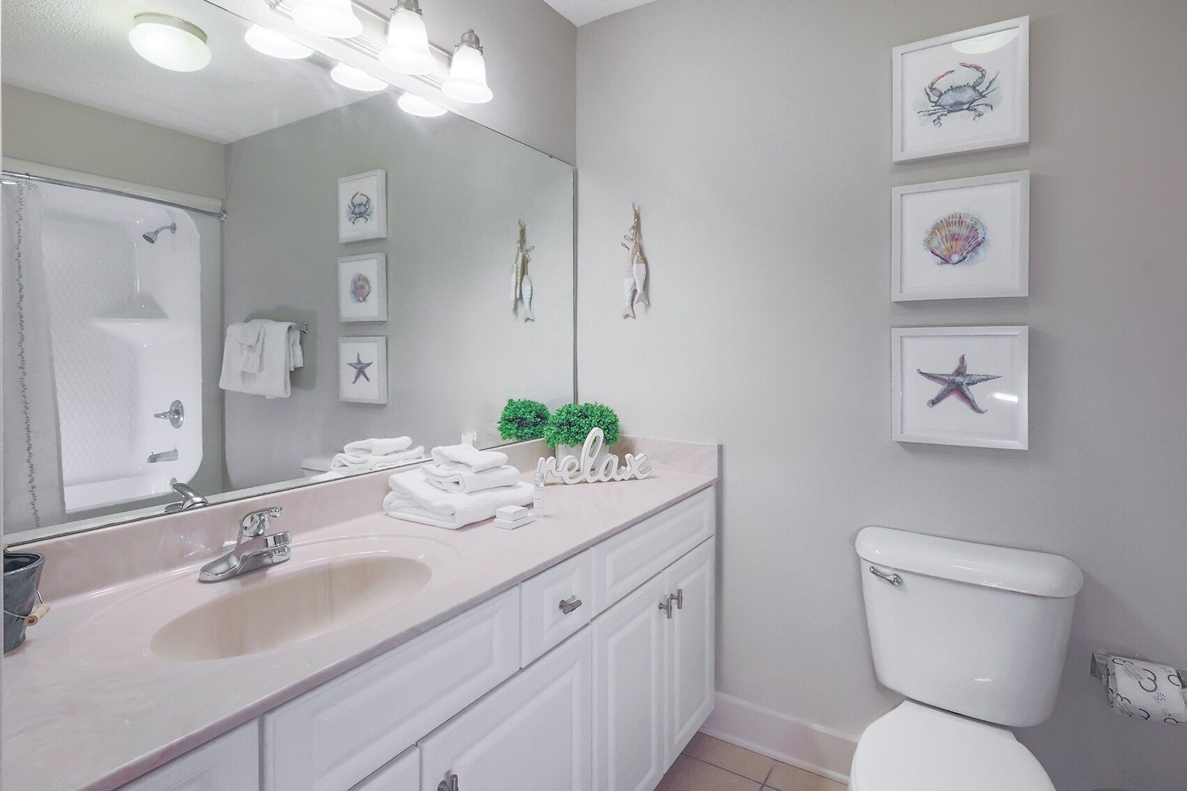 A bathroom with a sink, mirror, and toilet. There are towels stacked on the counter, a small plant, and framed pictures on the wall.