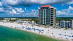 A high-rise hotel with a red roof, located on a beach with numerous umbrellas and a clear view of the ocean. - Phoenix West 1606 3 Bedroom Condo (Orange Beach)