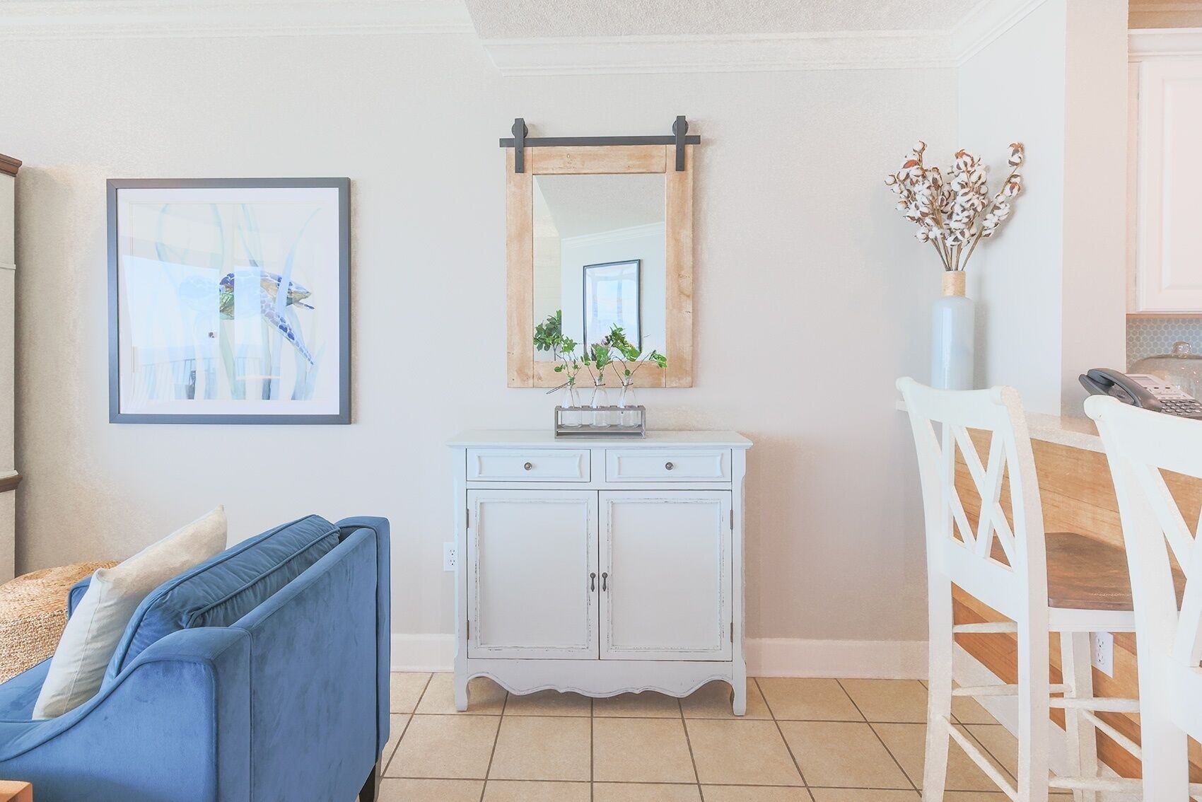 A living room with a blue sofa, a white cabinet, a framed picture, a mirror, and a vase with dried flowers.
