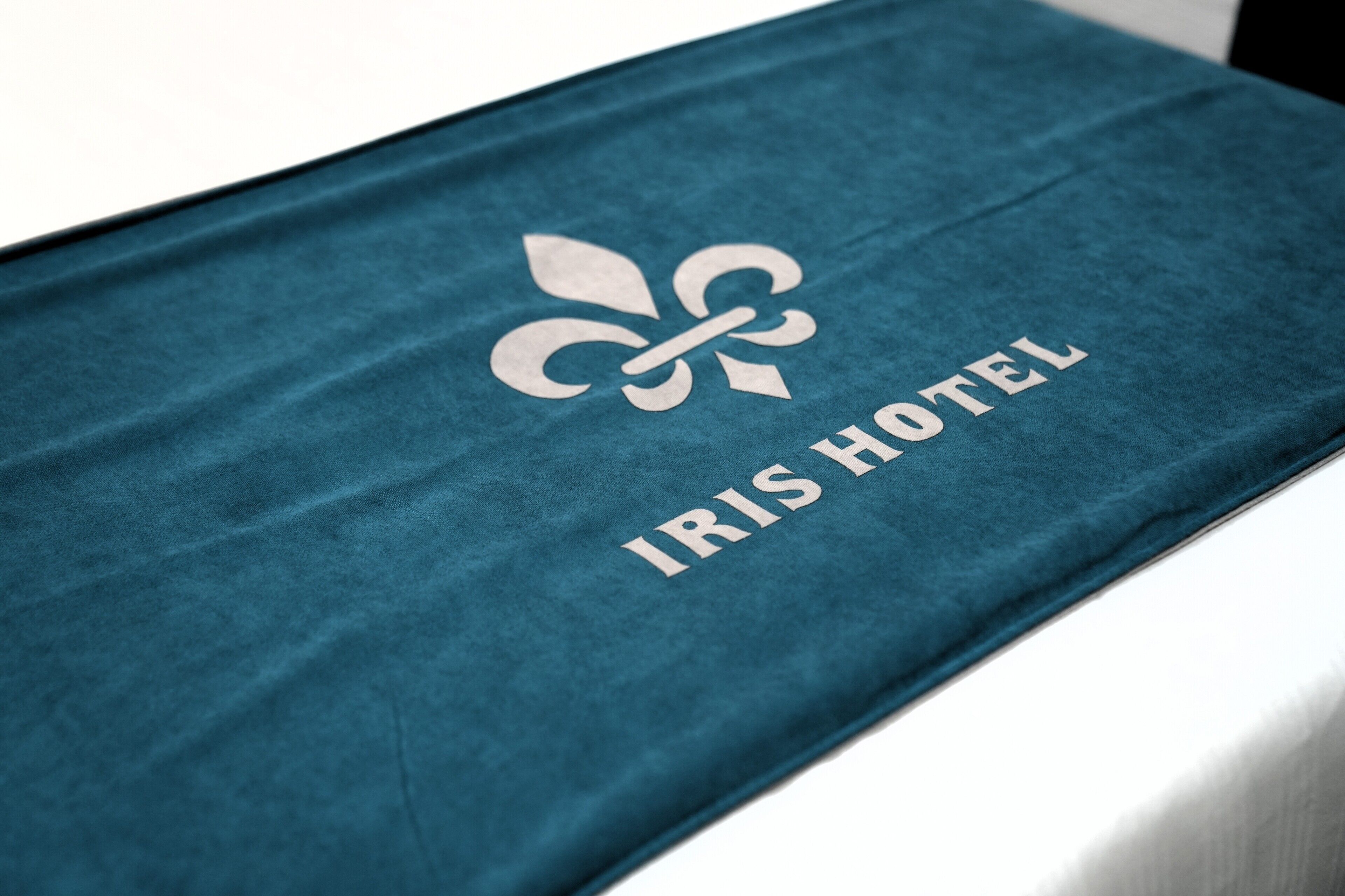 Una colcha azul aguamarina con un logo de fleur-de-lis y el texto 'IRIS HOTEL'.