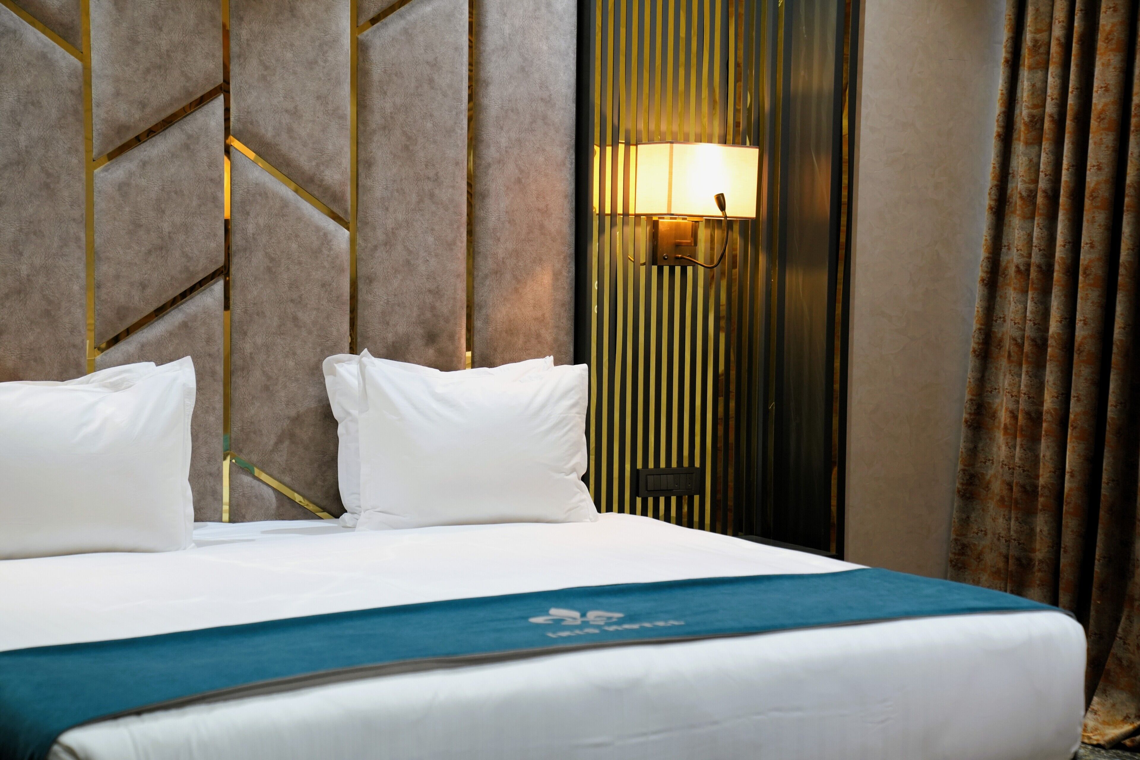 Quarto de hotel com cama, almofadas, colcha azul, luminária na parede e cortinas.