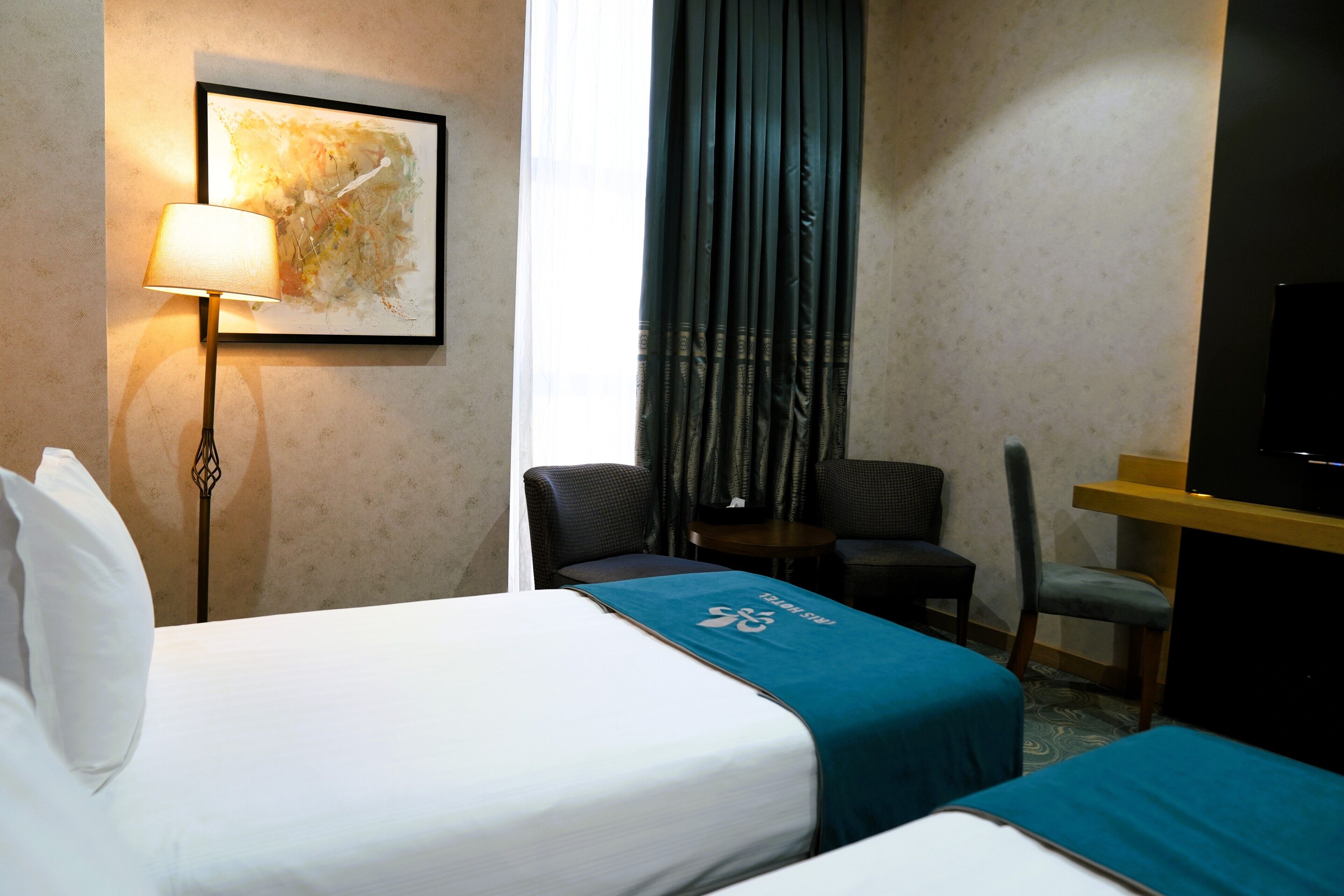 A hotel room with two beds, a desk, a chair, a lamp, and a television.