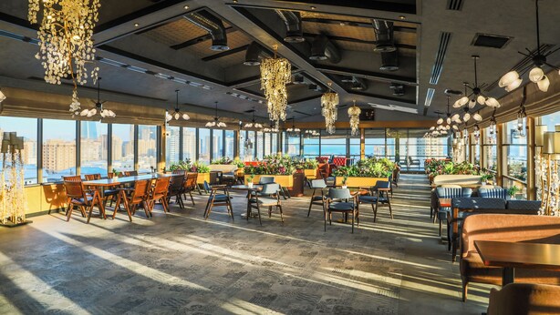 Un vaste espace intérieur doté de nombreuses tables et chaises, de grandes fenêtres offrant une vue sur la ville et de luminaires décoratifs suspendus.