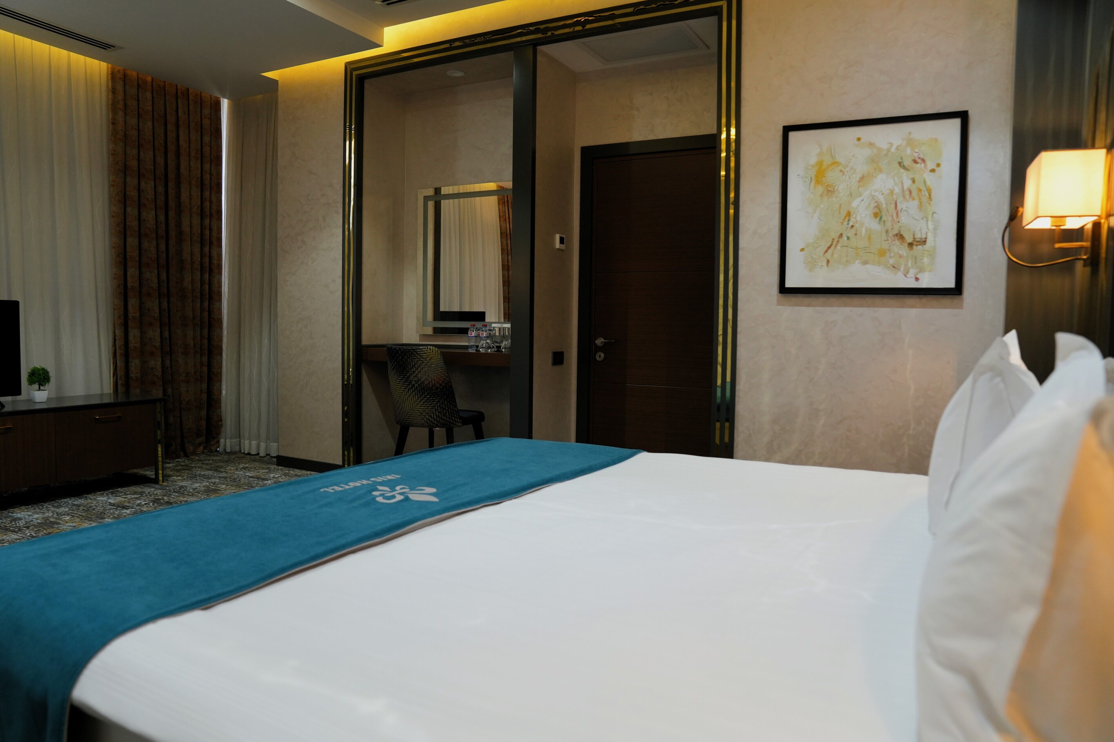 A hotel room with a bed, a desk, a chair, a television, a mirror, a door, and a wall-mounted light fixture.