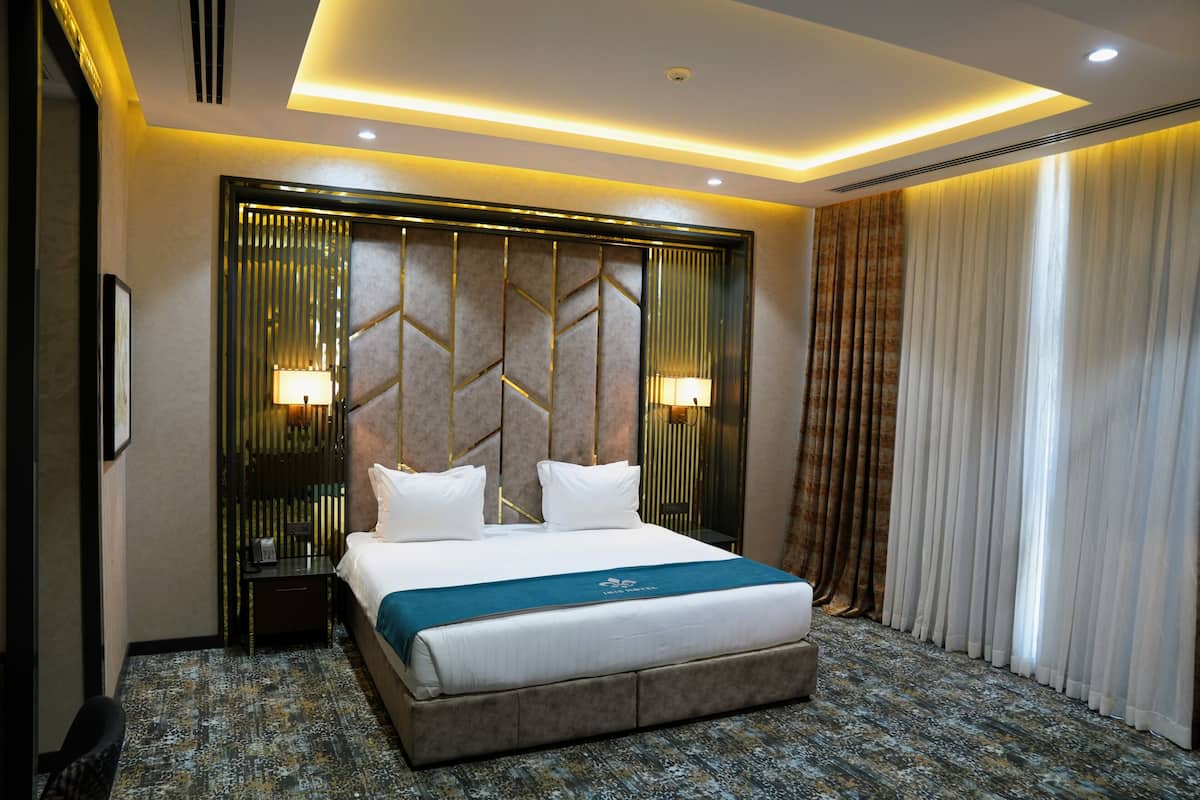 Habitación de hotel con una cama grande, cabecera con diseños geométricos, dos lámparas de noche, una mesita de noche y vistas al exterior a través de cortinas.