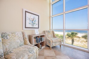A room with two patterned armchairs, a bookshelf, and a large window overlooking a beach. - Phoenix Gulf Shores 1903-1 1 Bedroom Condo (Gulf Shores)