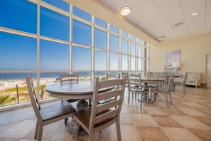 A dining area with large windows overlooking a beach, featuring round tables and wooden chairs.