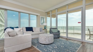 A spacious living area with a large window overlooking a beach, a sofa, a patterned ottoman, and a chair. - Phoenix Orange Beach Ii 405 4 Bedroom Condo (Orange Beach)