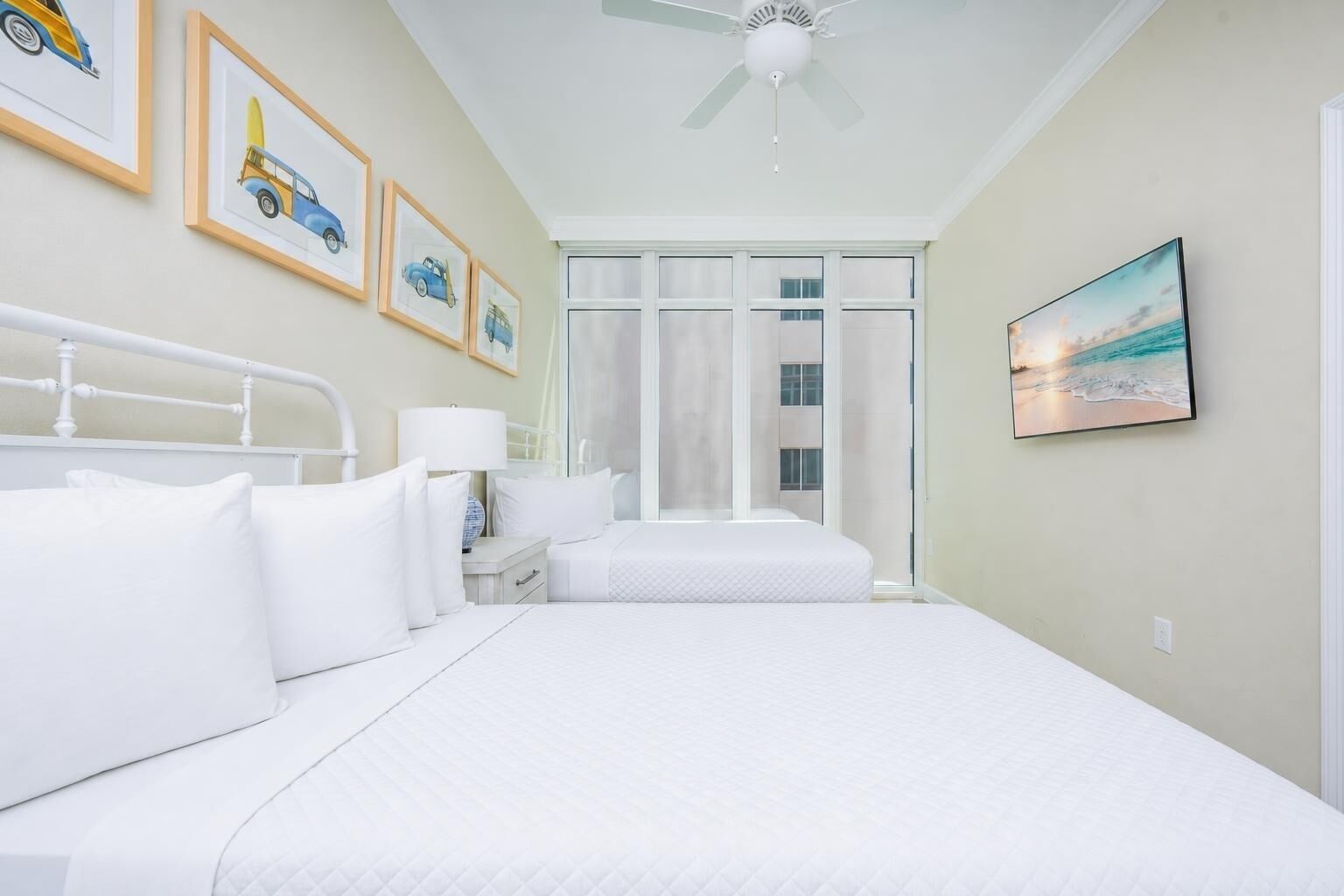 Un dormitorio con una cama, una mesita de noche, una lámpara, un ventilador de techo, una ventana con cortinas y cuadros en la pared.