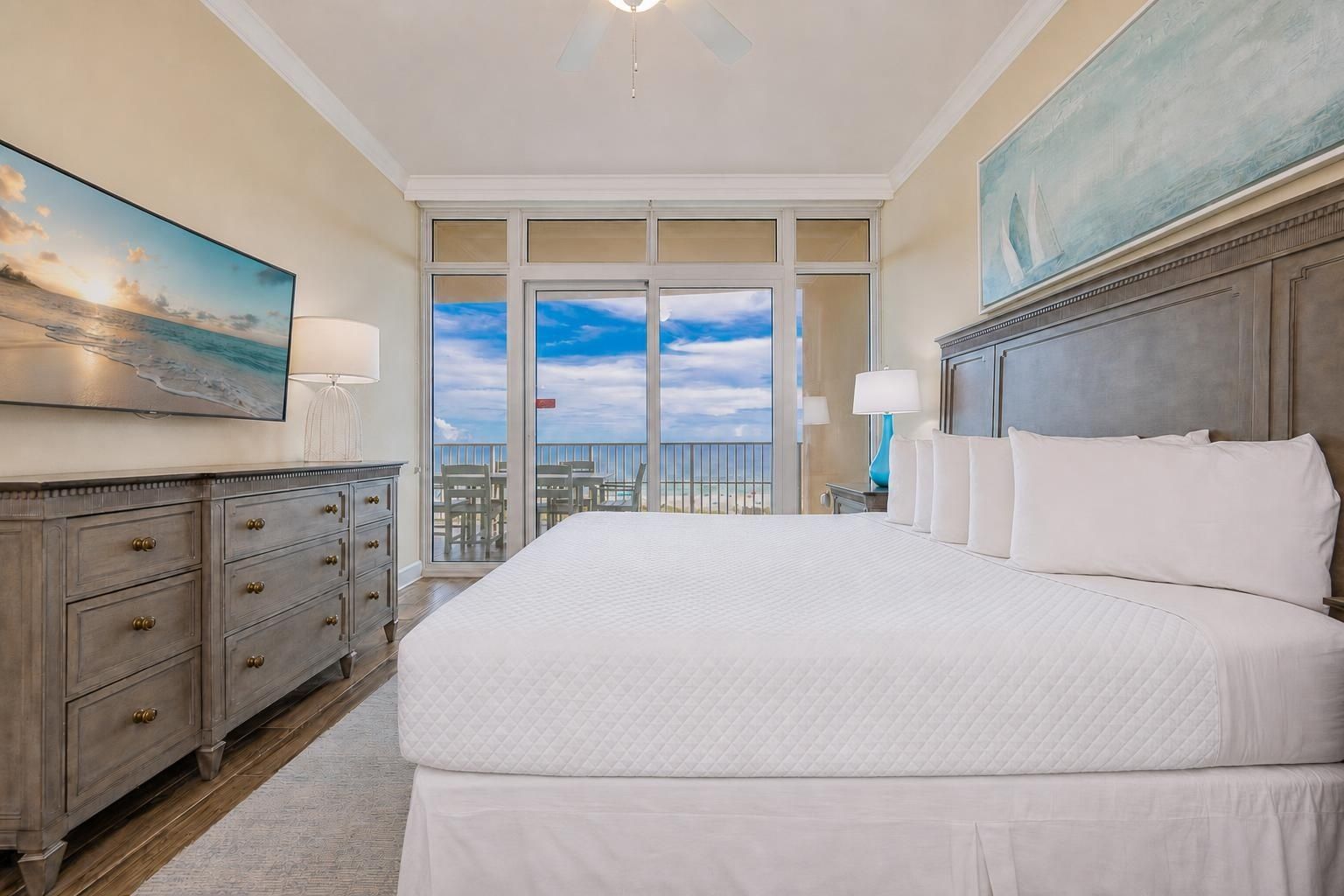 A hotel room with a large bed, a dresser, a painting, and a view of the beach through the balcony doors.