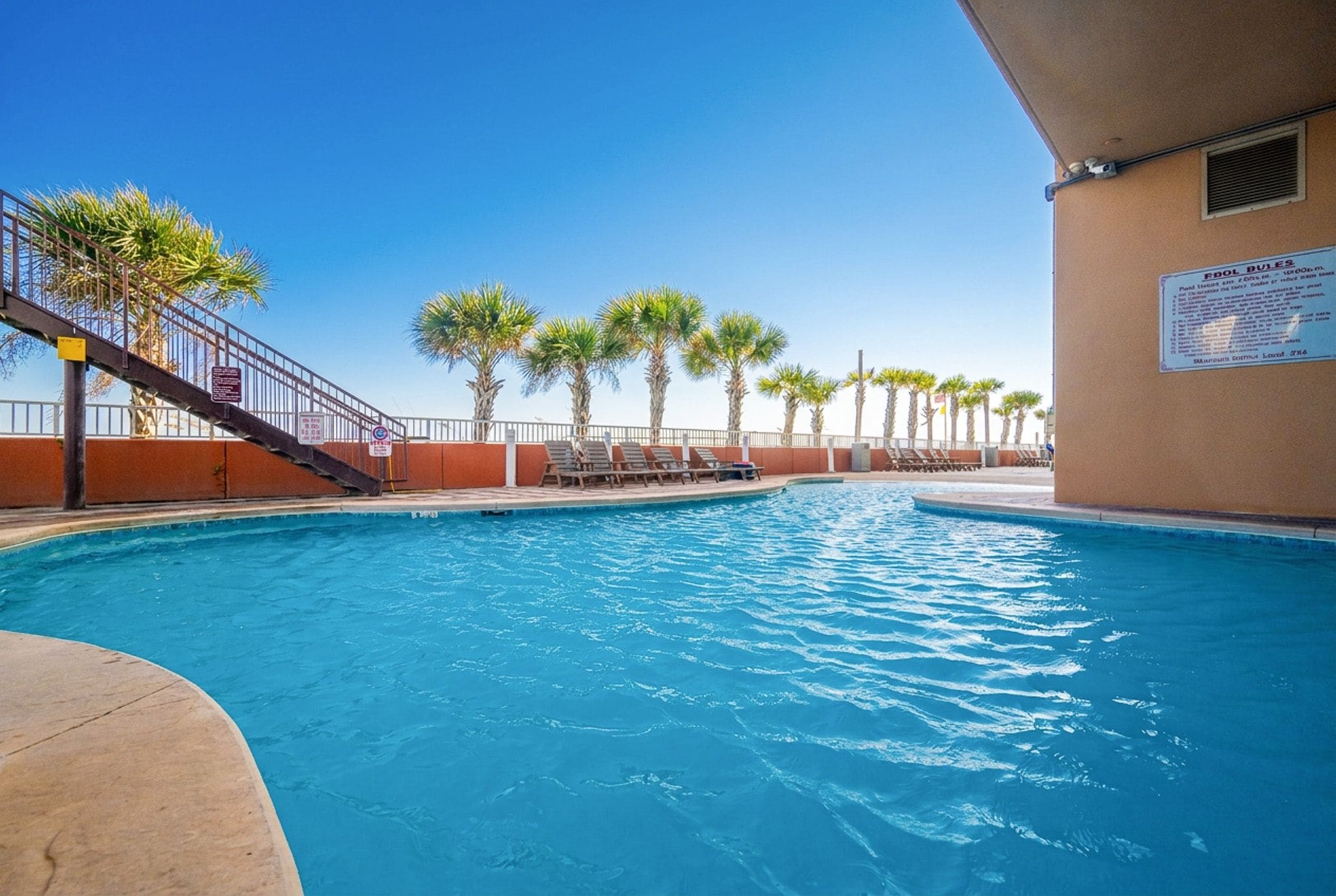 A swimming pool with a clear blue water surface, surrounded by palm trees and a fenced area with lounge chairs.