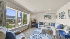 A spacious living room with a large window offering a view of a residential area and hills. There is a sofa, a smaller couch, and a blue patterned rug.