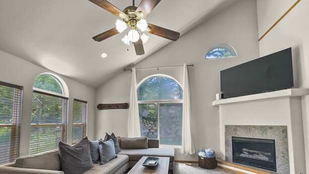 A spacious living room with a large sofa, a coffee table, and a fireplace. There are large windows and a ceiling fan.