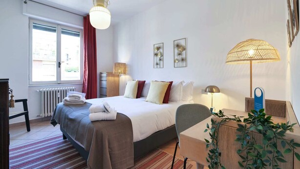 Ein Hotelzimmer mit Bett, Schreibtisch, Stuhl, Heizkörper, einem Fenster mit Vorhängen und Wandbildern.