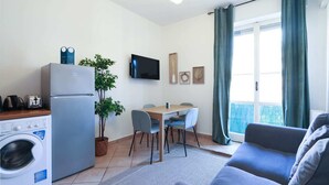 Ein kompakter Wohnraum mit Küchenzeile, Essbereich, einer Couch und einem großen Fenster mit Vorhängen.