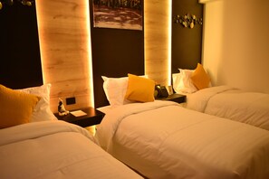 A hotel room with two beds, each with white bedding and a single yellow pillow. There is a nightstand with a phone and a small statue on each bed. The room has a wooden headboard and a painting on the wall.