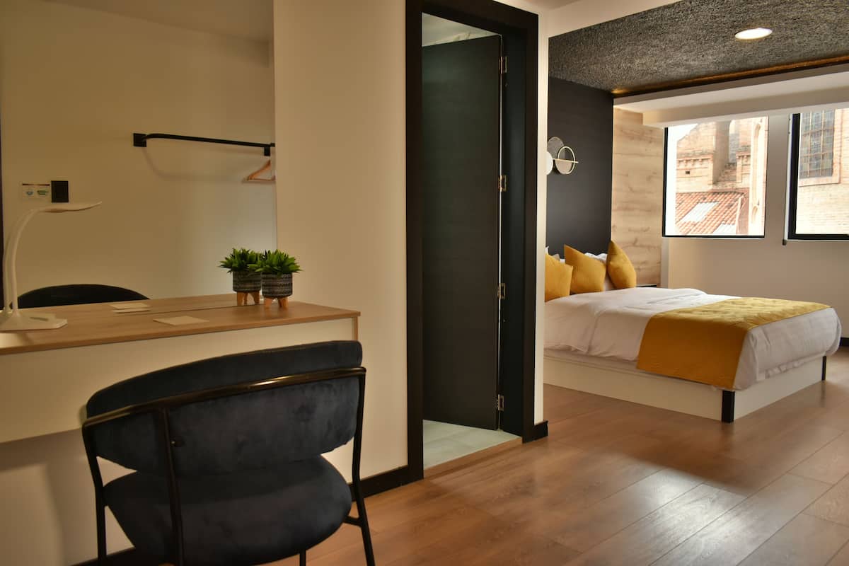 Una habitación de hotel moderna con un escritorio de madera, una silla azul oscuro, una cama con ropa de cama blanca y un plaid amarillo, y vistas de un edificio a través de una ventana.