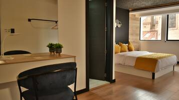 A modern hotel room with a wooden desk, a dark blue chair, a bed with white bedding and a yellow throw, and a view of a building through a window.
