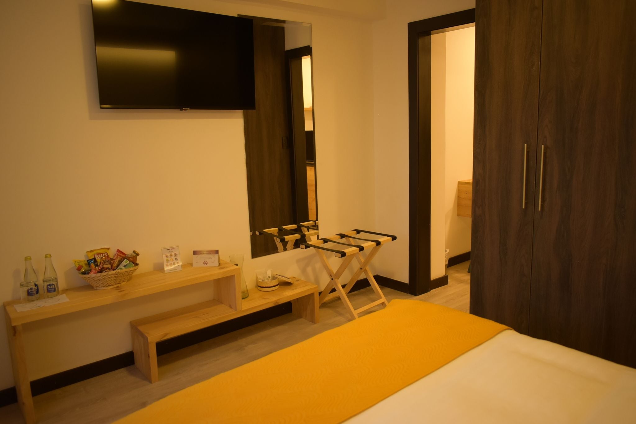 A hotel room with a bed, a wooden bench, a small table with snacks, a TV mounted on the wall, and a wardrobe.
