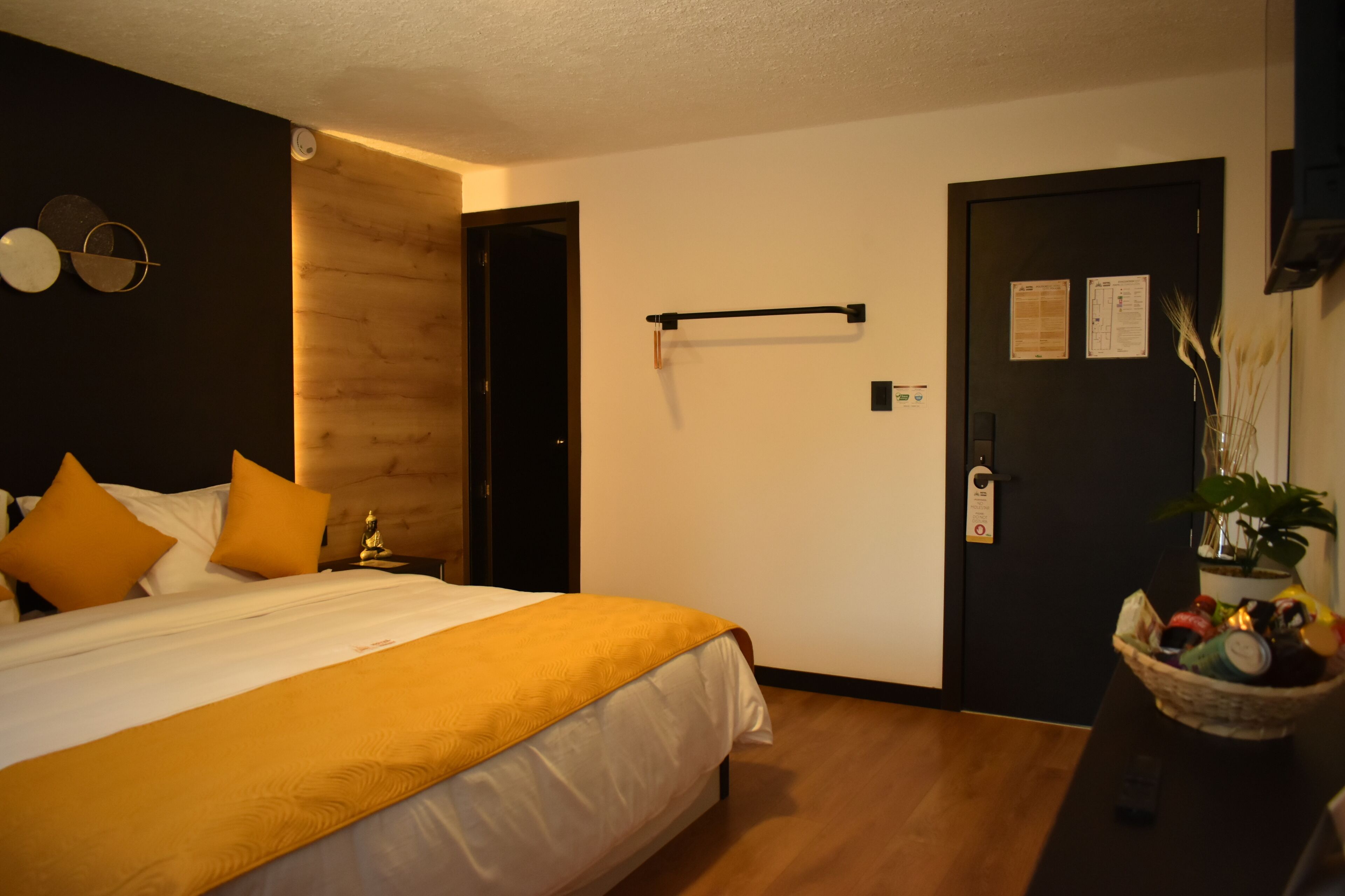 Une chambre d’hôtel avec un lit, une tête de lit en bois, une porte noire et une petite table sur laquelle se trouve une plante.