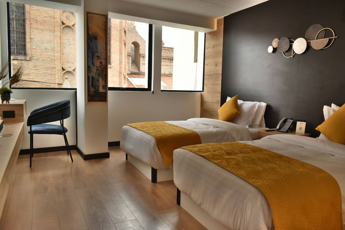 Habitación de hotel con dos camas, un escritorio y una silla. Amplias ventanas con vista a un edificio de ladrillo.