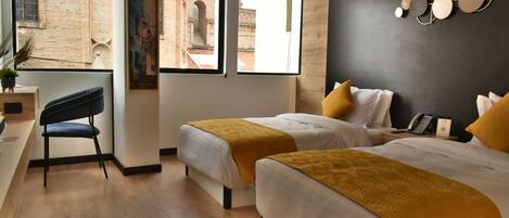A hotel room with two beds, a desk, and a chair. There are large windows with a view of a brick building.