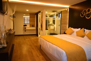 A modern hotel room with a large bed, a bathroom with a glass shower, and a wooden floor.
