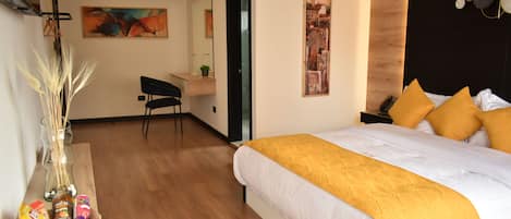 A modern hotel room with a large bed, a desk, and a chair. There is a basket of snacks on the desk and a vase with dried plants.