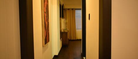 Un couloir étroit avec un plancher en bois, un tableau accroché au mur de gauche, un miroir et une fenêtre avec des rideaux au fond.