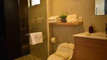 A modern bathroom with a glass shower enclosure, a toilet, a sink with a soap dispenser, and a shelf with towels and a potted plant.