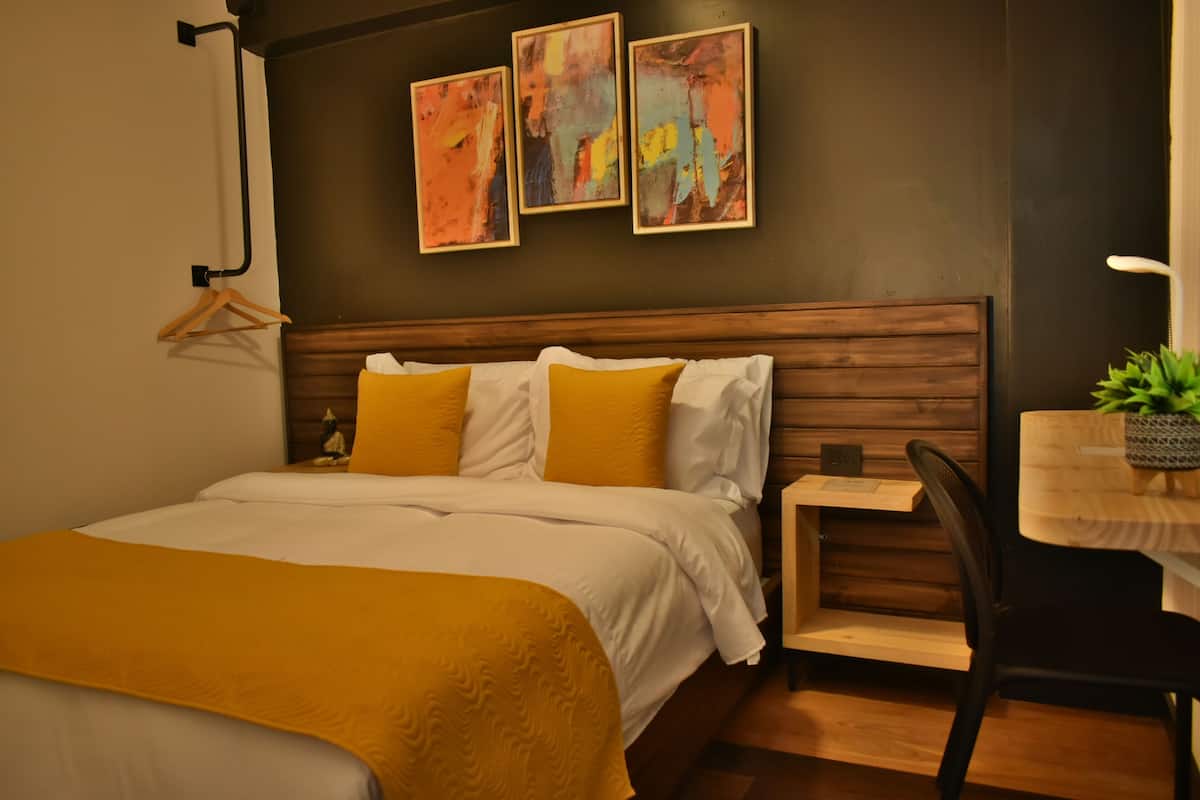 Una habitación de hotel moderna con cabecera de madera, cama con ropa de cama blanca y mostaza, un escritorio pequeño con una silla y arte abstracto en la pared.