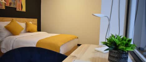 A hotel room with a bed, a desk, a chair, a potted plant, and artwork on the wall.