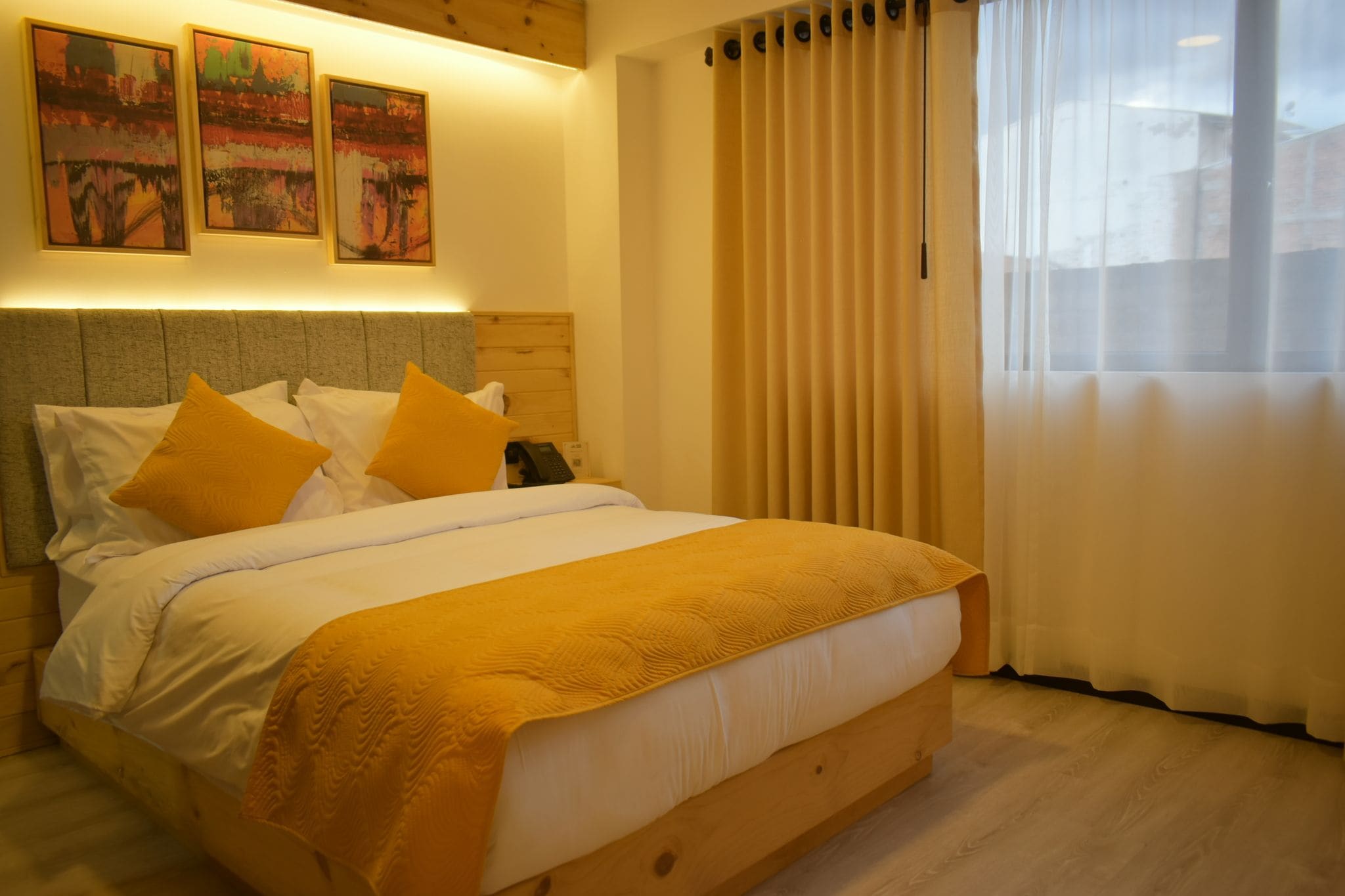 A hotel room with a bed, a wooden headboard, a bedside table with a phone, and a window with curtains.