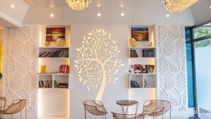 Interior moderno com paredes brancas, padrões decorativos de folhas e um grande mural de árvore. Há estantes, uma mesa redonda e duas cadeiras.