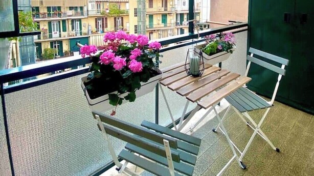 Een balkon met een tafel en stoelen, een potplant met roze bloemen en uitzicht op gebouwen.
