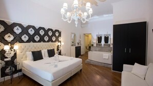A bedroom with a large bed, a chandelier, a wardrobe, and a bathroom in the background. - Navona Vita Suites (Rome)