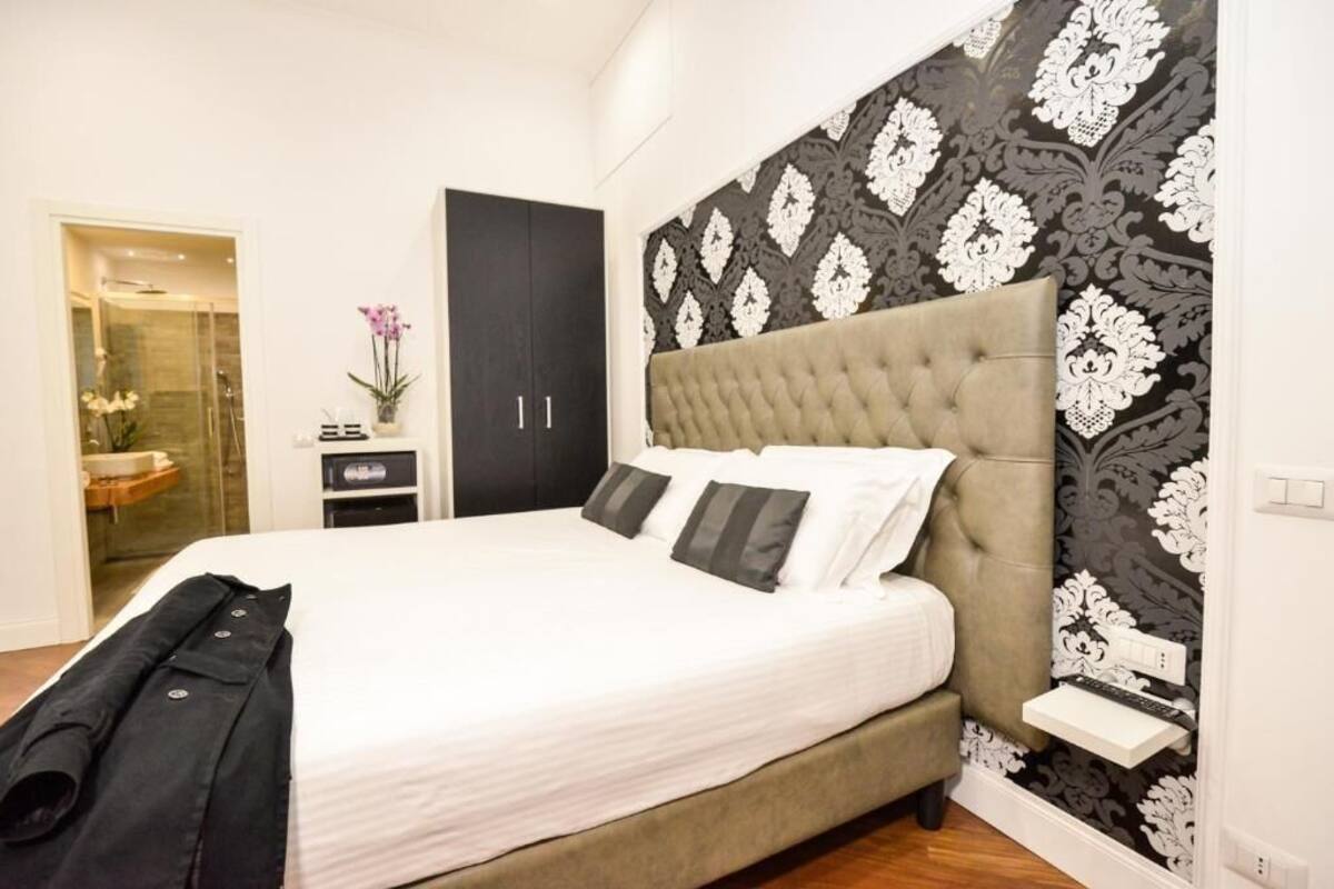 A bedroom with a large bed, a dark wardrobe, a small bedside table, and a shower area visible through a glass door.