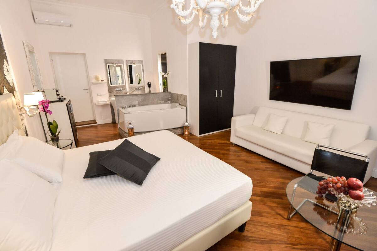 A modern hotel room with a large bed, a white sofa, a TV, and a bathroom with a bathtub and a large mirror.