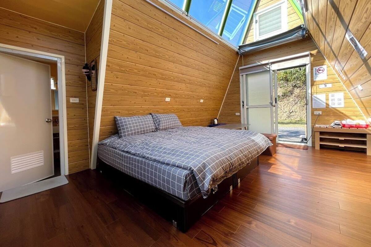 A bedroom with a wooden sloped ceiling, a bed with a checkered bedspread, and a view of the outdoors through a glass door.