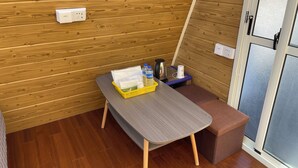 Quarto compacto com parede de madeira, uma mesa cinza com uma cesta amarela, um banco marrom e uma porta com painel de vidro fosco.