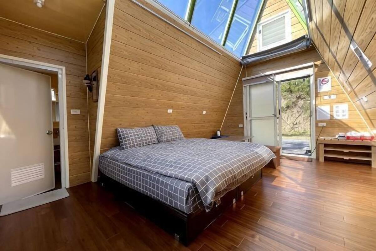 A bedroom with a wooden floor, a bed with a checkered bedspread, a skylight, and a door leading outside.