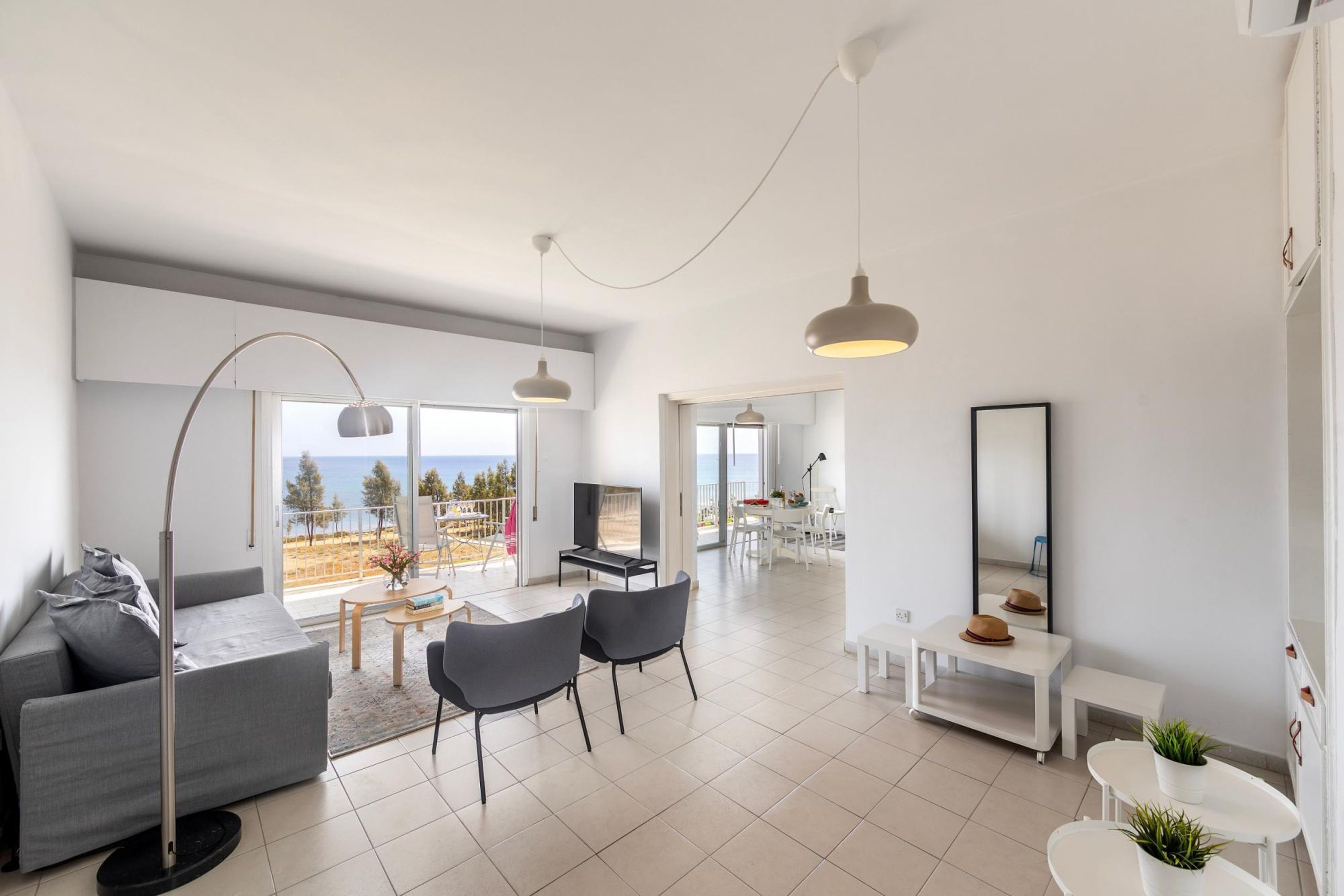 A spacious living area with a sofa, chairs, a coffee table, and a TV. There is a balcony with a view of the sea.