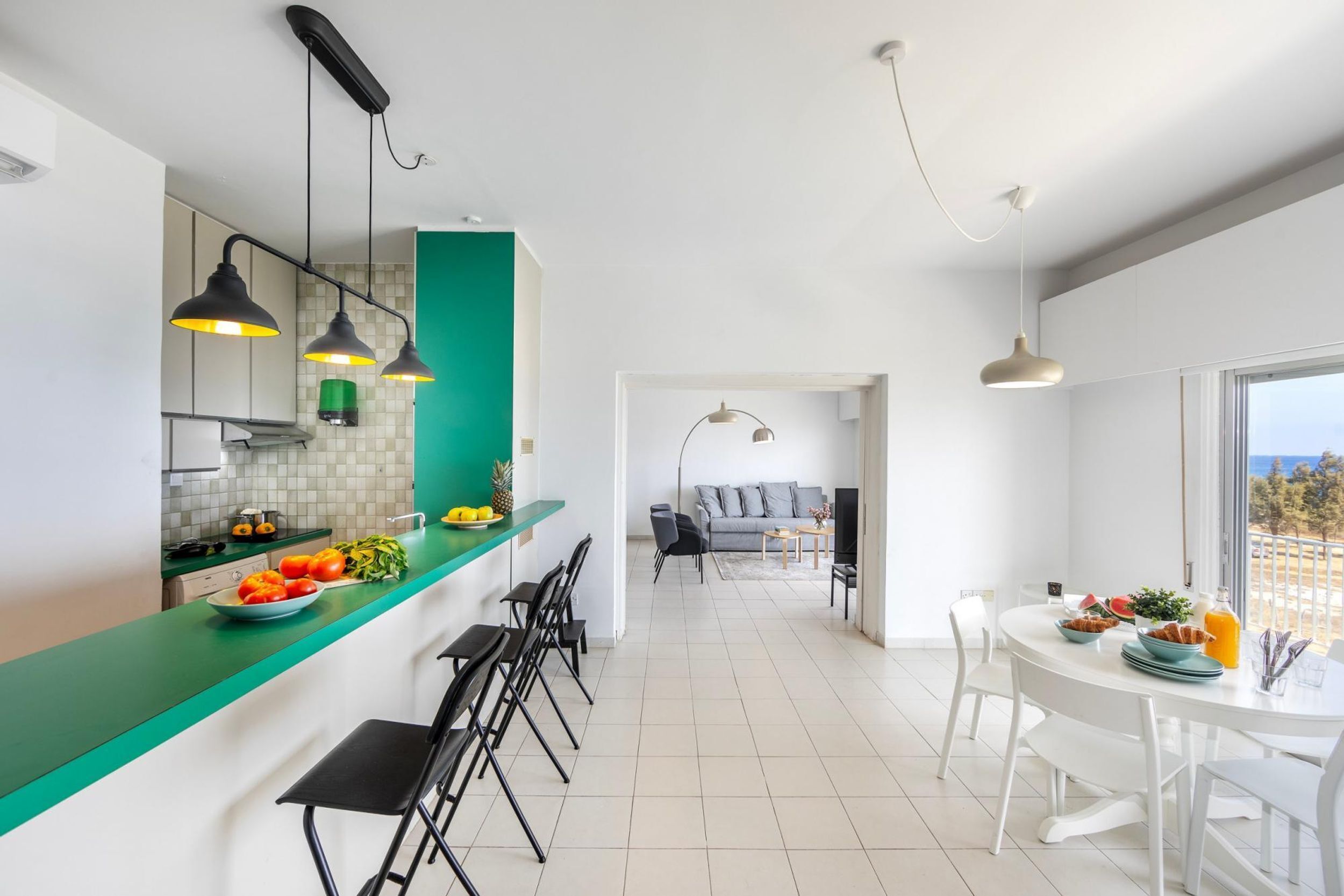 Une cuisine moderne avec un plan de travail vert, des tabourets de bar noirs et des luminaires suspendus. On aperçoit à l’arrière-plan un coin repas avec une table et des chaises blanches.
