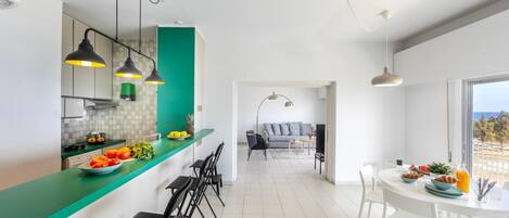 Une cuisine moderne avec un plan de travail vert, des tabourets de bar noirs et des luminaires suspendus. On aperçoit à l’arrière-plan un coin repas avec une table et des chaises blanches.