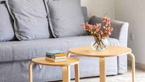 Un salon avec un canapé gris, une petite table ronde en bois sur laquelle est posé un vase de fleurs, et une pile de livres sur le canapé.