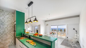 Une cuisine moderne avec un plan de travail vert, un évier et un éclairage au plafond. On y trouve également un coin repas avec une table et des chaises, et une vue sur l’océan grâce à de grandes fenêtres.